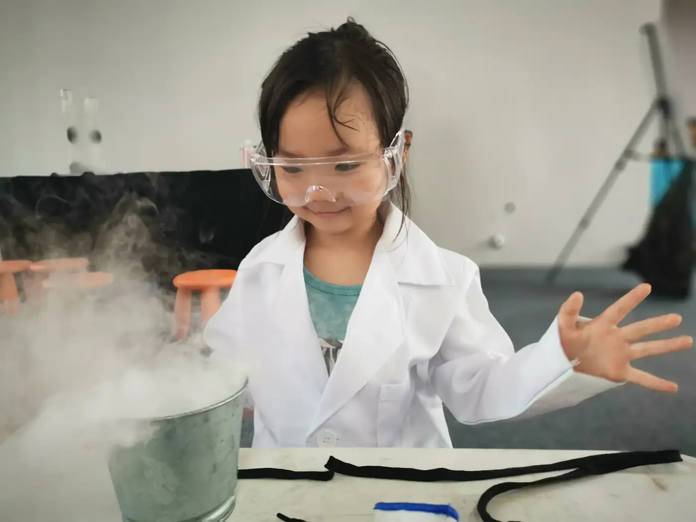 A girl in a lab coat and goggles watching vapour rise from an experiment