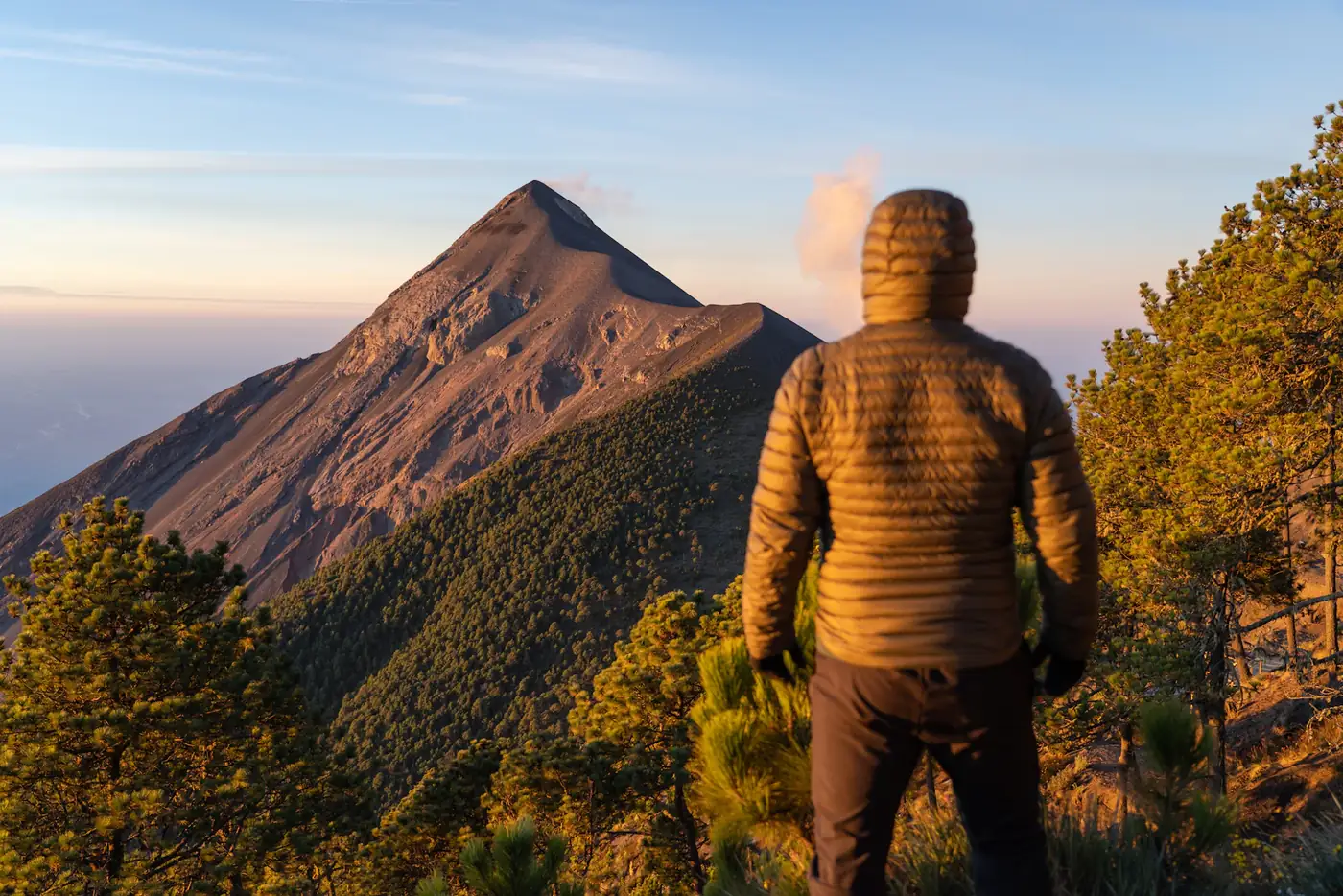 A hiker in an orange jacket looking up at a volcano peak